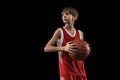 Cropped portrait of young boy, basketball player in red uniform standing, posing with ball isolated over black Royalty Free Stock Photo