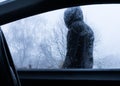 Looking through a car window at a mysterious scary hooded figure standing outside. On a rainy winters day