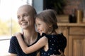 Young hairless mother standing by window embracing little daughter