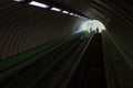 Looking up through a dark inclined escalator tunnel in subway undergraound setting towards bright entrance with small figures of p