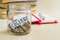 A lot of coins and college word in a glass jar near a white ballpoint pen and few books on a table. Saving money for college and Royalty Free Stock Photo