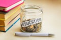 A lot of coins and education word in a glass jar near a white ballpoint pen and three books on a table. Saving money for education Royalty Free Stock Photo