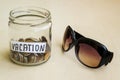 A lot of coins and vacation word in a glass jar near sunglasses on a table. Saving money for vacation concept Royalty Free Stock Photo