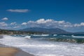 Lots of white waves foam on sandy sea beach, blue water and blue sky with white clouds around the mountain in distance. Royalty Free Stock Photo