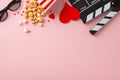 Top view snapshot of clapperboard, 3D glasses, striped popcorn container, heart-shaped ornaments, marshmallows on pink surface