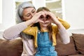 Love. Portrait of little granddaughter showing heart sign together with her loving grandmother while having fun at home