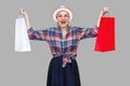 Love shopping! Satisfied modern grandma in white hat and in checkered shirt holding shopping bags and triumphing with raised arms