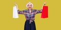 Love shopping! Satisfied modern grandma in white hat and in checkered shirt holding shopping bags and triumphing with raised arms