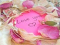 Love you message on pink sticky note with dry rose and orchid flower petals and jewelry ring and chain on wooden background Royalty Free Stock Photo