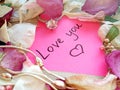 Love you message on pink sticky note with dry rose and orchid flower petals and silver jewelry ring and chain on wooden back Royalty Free Stock Photo