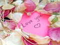 Love you message on pink sticky note with dry rose and orchid flower petals and silver jewelry ring and chain on wooden back Royalty Free Stock Photo