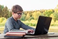 Loveable boy in glasses sitting at table, smiling while looking at monitor of laptop. Pupil enjoy studying in park.