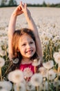 Loveable female child with bright smile standing surrounded by white fluffy dandelions in field and stretching arms up.