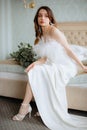 Loveable lady sit on bed in shoulderless dress adorned with feathers and heels. Bride with tassel earrings look aside.