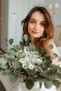 Loveable young bride with dark hair and brown eyes sitting on bed, holding bouquet of white flowers and smiling softly. Royalty Free Stock Photo