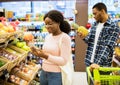 Lovely black woman and her boyfriend shopping for fruits, checking grocery list on mobile phone at supermarket Royalty Free Stock Photo