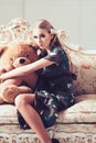 Lovely blond girl hugging huge teddy bear while sitting on retro sofa. Slim woman in flowery gray dress in her room