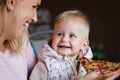 Lovely blond little baby girl in beautiful white dress biting on piece of tasty pizza