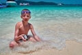 Lovely boy with swimming goggles sits in the turquoise water of the tropical sea surf line.