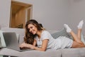 Lovely brunette girl in white socks using laptop with charming smile. Pleased young lady posing on gray couch with Royalty Free Stock Photo