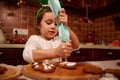 Lovely child girl, little baker confectioner squeezing glaze from baking bag, on fresh gingerbread in Christmas interior