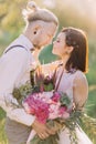 The lovely close-up portrait of the newlyweds close to each other and holding the pink bouquet in the sunny forest.