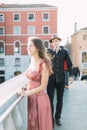 Lovely couple in Venice. Pretty young girl in long rose dress standing on the bridge of venetian canal, young man in black clothes Royalty Free Stock Photo