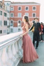 Lovely couple in Venice. Pretty young girl in long rose dress standing on the bridge of venetian canal, young man in black clothes Royalty Free Stock Photo