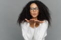 Lovely curly woman sends air kiss, has lips folded, keeps palms outstretched, wears transparent glasses, white jumper, expresses
