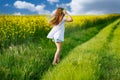 Teenage girl in white dress and Ukrainian wreath runs through yellow fields and green meadows, against cloudy sky Royalty Free Stock Photo