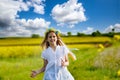 Teenage girl in white dress and Ukrainian wreath runs through yellow fields and green meadows, against cloudy sky Royalty Free Stock Photo