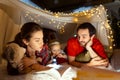 Lovely family, mother, father and daughter lying inside self-made hut, tent in room in the evening and reading book Royalty Free Stock Photo