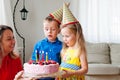 Lovely girl and boy twins in party hats makes a wish before blow out four candles on a birthday cake. Mom smiles and holds a cake. Royalty Free Stock Photo