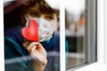 Lovely little school kid boy by a window wearing medical mask and holding wooden heart during pandemic coronavirus Royalty Free Stock Photo