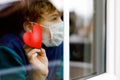 Lovely little school kid boy by a window wearing medical mask and holding wooden heart during pandemic coronavirus Royalty Free Stock Photo