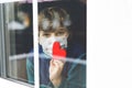 Lovely little school kid boy by a window wearing medical mask and holding wooden heart during pandemic coronavirus Royalty Free Stock Photo