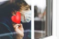Lovely little school kid boy by a window wearing medical mask and holding wooden heart during pandemic coronavirus Royalty Free Stock Photo