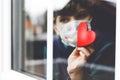 Lovely little school kid boy by a window wearing medical mask and holding wooden heart during pandemic coronavirus Royalty Free Stock Photo