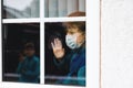 Lovely little school kid boy by a window wearing medical mask at home or school during pandemic coronavirus quarantine Royalty Free Stock Photo