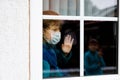 Lovely little school kid boy by a window wearing medical mask at home or school during pandemic coronavirus quarantine Royalty Free Stock Photo