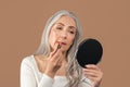 Lovely mature lady applying lipstick in front of mirror, taking care of herself on brown studio background Royalty Free Stock Photo