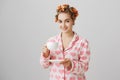 Lovely ordinary european woman in hair curlers and pyjamas, holding cup while drinking coffee or tea in morning, smiling