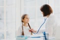 Lovely small girl listens consult of professional experienced doctor who listens her lungs with stethoscope, comes on medical