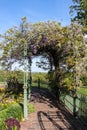 A lovely stroll down a sidewalk leading under metal arbor covered in wisteria with decorative fence and flowers and countryside in