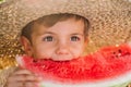 Lovely toddler boy eating fresh red watermelon. Yummy portrait of cheerful child in straw hat sits by the table in Royalty Free Stock Photo