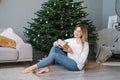 Lovely woman is sitting on the floor near the Christmas tree in the living room and holding a surprise gift in her hands Royalty Free Stock Photo