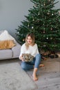 Lovely woman is sitting on the floor near the Christmas tree in the living room and holding a surprise gift in her hands Royalty Free Stock Photo