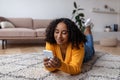 Young black lady lying on floor with cellphone, checking news, sending sms to friend, video chatting online from home Royalty Free Stock Photo