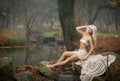 Lovely young lady sitting near river in enchanted woods. Sensual blonde with white clothes posing provocatively in autumnal park. Royalty Free Stock Photo