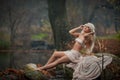 Lovely young lady sitting near river in enchanted woods. Sensual blonde with white clothes posing provocatively in autumnal park. Royalty Free Stock Photo